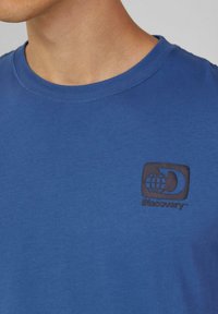 Niebieska bawełniana koszulka z okrągłym dekoltem i czarnym, wytłoczonym logo z motywem globusa po lewej stronie klatki piersiowej.