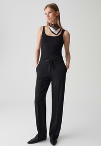 Canottiera nera e pantaloni neri con vita regolabile. Il modello indossa una sciarpa a motivi attorno al collo, in piedi contro uno sfondo chiaro.