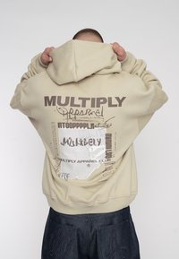 Béžová mikina s kapucí a velkým grafickým potiskem na zádech, který obsahuje text a vzory. Má stahovací kapuci a žebrované manžety.