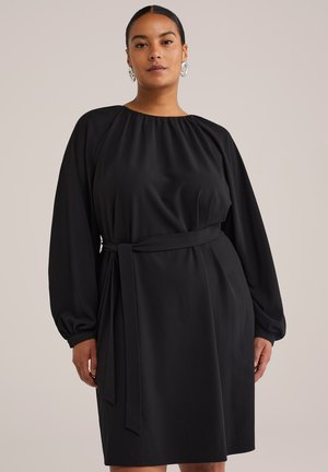 Vestido negro con escote fruncido, mangas largas y abullonadas, y cinturilla ceñida. Confeccionado en tejido liso, con un corte a la altura de la rodilla y de color sólido.