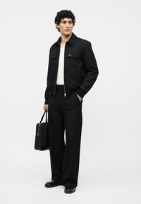 Uomo con giacca nera, pantaloni neri larghi, camicia bianca, scarpe nere, che tiene una valigetta nera, in piedi contro uno sfondo bianco.