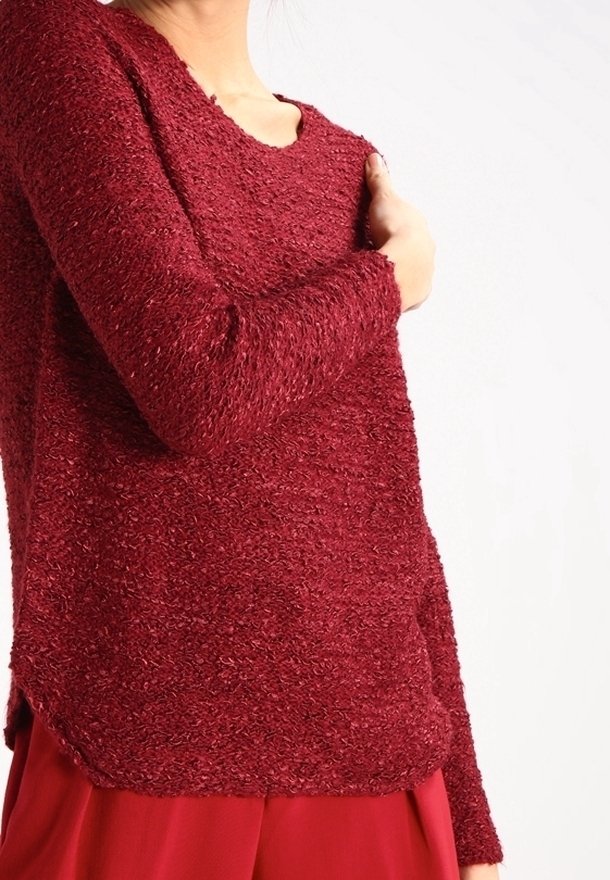 Femme portant un pull bordeaux texturé à manches longues et un pantalon ample assorti, une main touchant son épaule.