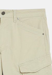 Beige cargo pants with front slant pocket, metal button closure, belt loop, and flap pocket on side leg.