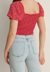 Rotes crop Top mit floralem Muster, kurzen Puffärmeln und gerafftem Rücken; kombiniert mit hellblauen, hoch taillierten Jeans.