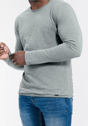 Homme portant une chemise à manches longues gris clair et un jean bleu ajustant sa manche gauche sur un fond blanc uni.