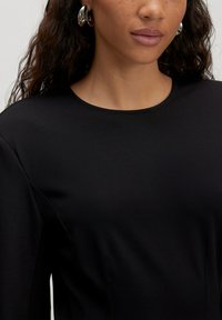 Vrouw met krullend haar die zilveren hoepelringen draagt en een zwarte ronde hals top, waarbij haar nek en bovenborst zichtbaar zijn.