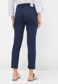 Navyblauwe slim fit broek van glad materiaal, met achterzakken en een middelhoge taille. Schoenen zijn lichtkleurig met een gespsluiting.