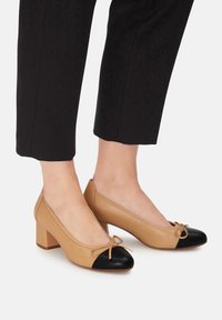 Zapato de tacón de cuero beige con puntera negra, que presenta un lazo en la parte frontal y un tacón cuadrado. Combinado con pantalones negros cortos.