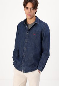 Denim shirt in dark blue with a button-up front, collar, and red logo detail. Model wears a grey t-shirt underneath. Smooth texture, relaxed fit.