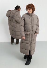Abrigo largo gris tipo plumas con cremallera, bolsillos laterales y un cuello alto. Diseño acolchado con una textura suave y brillante.