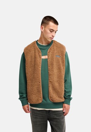 Junger Mann trägt eine braune Fleeceweste über einem grünen Sweatshirt und dunklen Jeans, schaut mit neutralem Gesichtsausdruck nach unten.