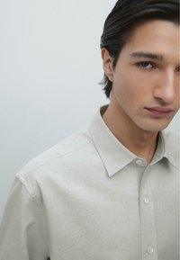 Camicia grigia chiaro con bottoni e colletto strutturato, caratterizzata da una texture sottile e un taglio dritto, parzialmente mostrata su uno sfondo neutro.