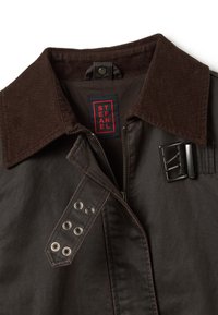 Veste marron avec col en velours côtelé, fermeture éclair à l'avant, détail de boucle et œillets en métal sur les épaules. L'étiquette indique "STEFAN." Texture lisse.