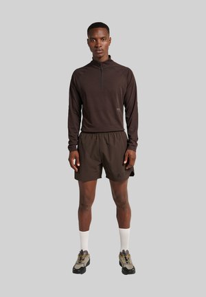 Donkerbruin T-shirt met lange mouwen en een ritssluiting bij de kraag, gecombineerd met bijpassende shorts. Het model draagt witte sokken en grijsbruin sportchoenen.