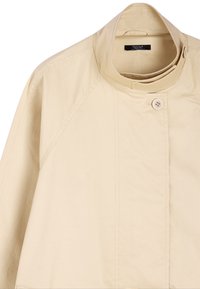 Beżowa, lekka kurtka z wysokim kołnierzem, z zapięciem na jeden guzik i rękawami raglanowymi. Materiał ma gładką fakturę.