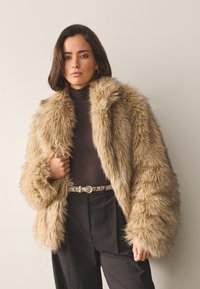 Faux fur jacket in light brown with a wide collar and oversized sleeves, paired with a dark turtleneck and plaid trousers. Textured and plush.