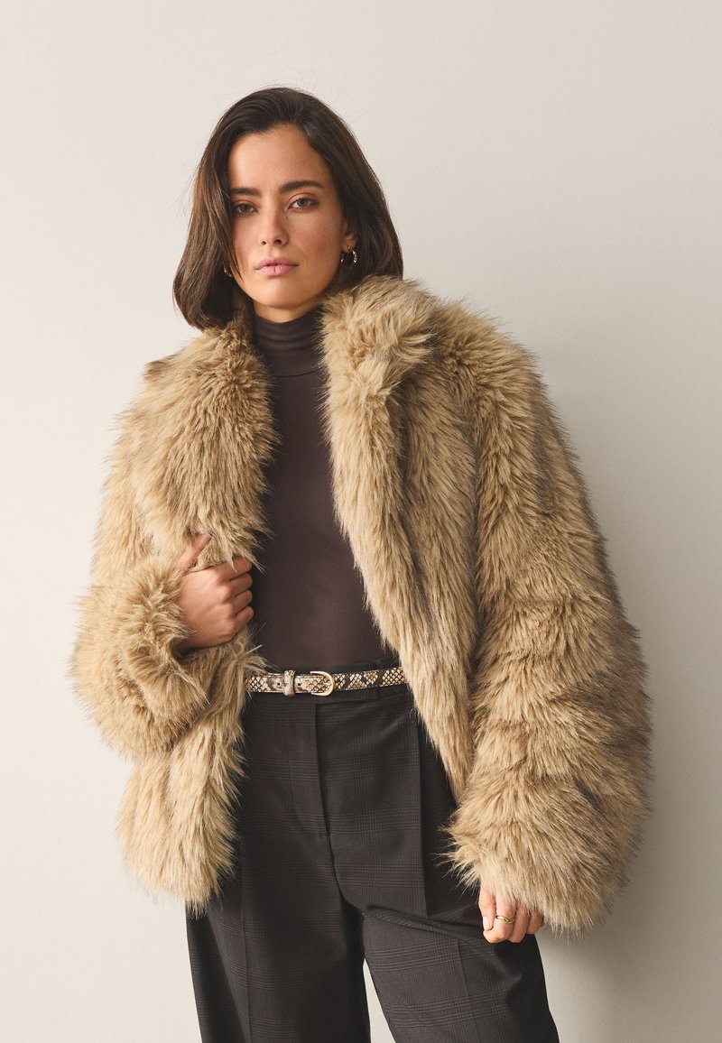 Faux fur jacket in light brown with a wide collar and oversized sleeves, paired with a dark turtleneck and plaid trousers. Textured and plush.