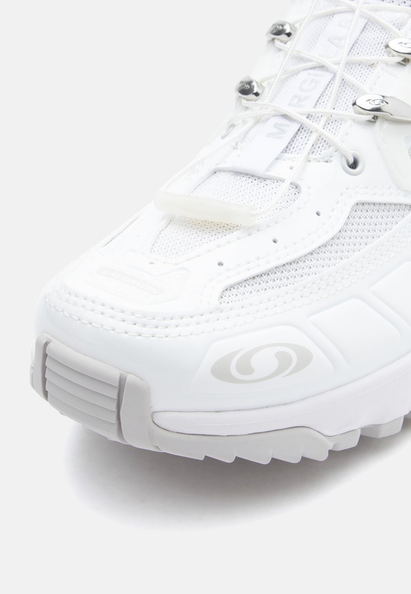 Weiße Sneaker mit Mesh- und synthetischen Materialien, runder Zehenpartie, gepolstertem Zungenbereich, metallischen Akzenten und strukturiertem Sohlenprofil mit Grip-Muster.