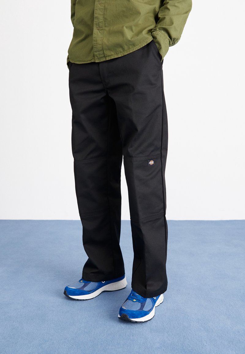 Homme portant un pantalon de travail noir, des baskets bleues et une chemise à manches longues vert olive, les mains dans les poches sur un sol bleu.