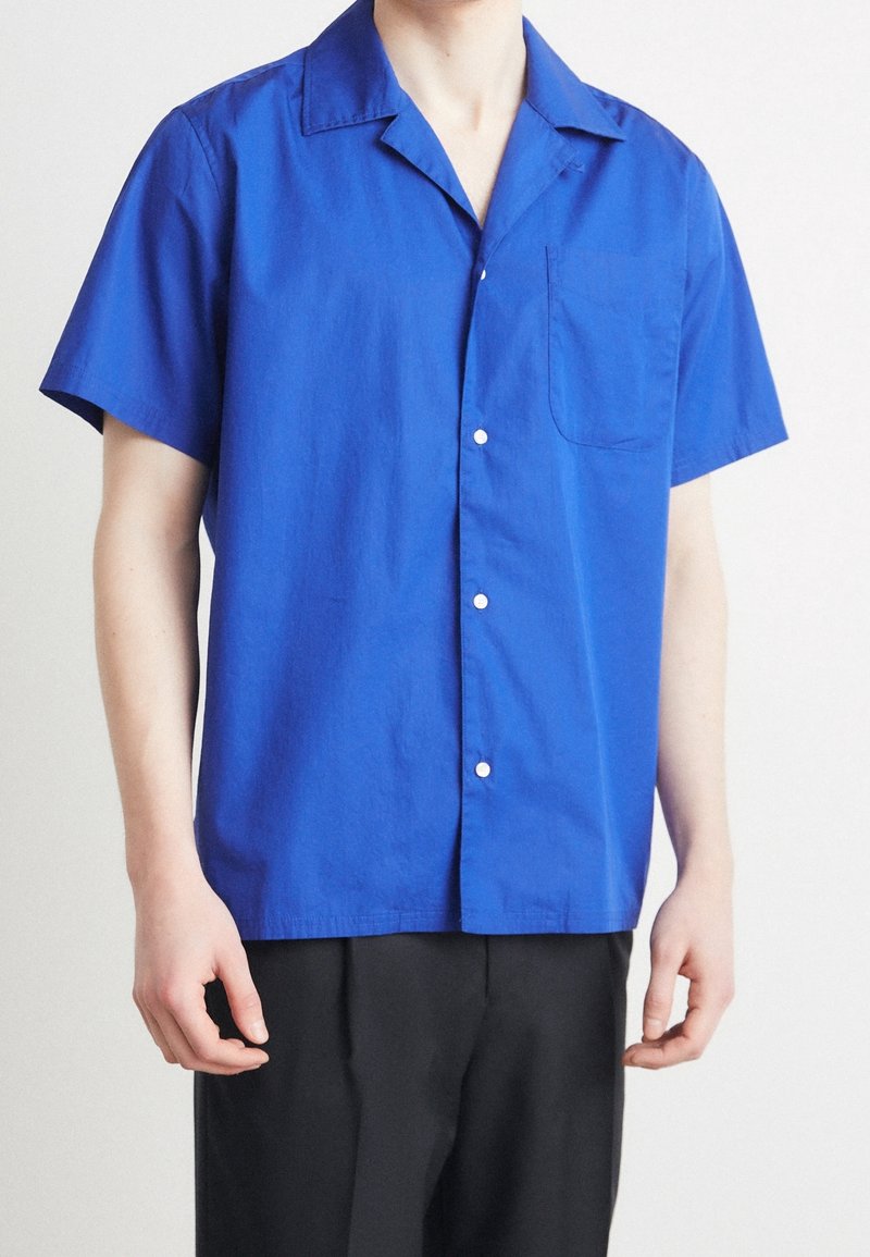Koszula z krótkim rękawem i kołnierzykiem, wykonana z jednolitego, jaskrawo niebieskiego materiału. Posiada kieszeń na piersi oraz zapięcie na białe guziki.