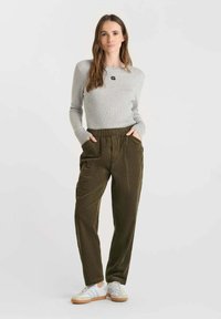 Woman standing with hands in pockets wearing a light gray sweater, olive green corduroy pants, and white sneakers on a plain background.