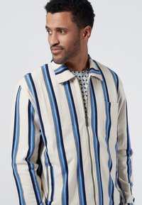 Randig zip-up jacka i lätt tyg; har blå och marinblå ränder på en krämfärgad botten, med krage och en bröstficka.