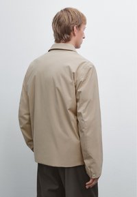 Giacca leggera beige con design a colletto, caratterizzata da maniche lunghe e una superficie liscia. La parte posteriore mostra una silhouette semplice e pulita.
