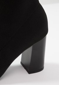 Gros plan sur une botte-socquette noire avec tige en tissu côtelé et talon épais, haut et bloc sur fond blanc.