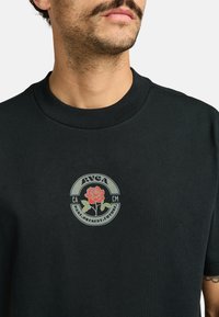 Camiseta negra de manga corta con cuello redondo que presenta un gráfico circular de una rosa y el texto "RVCA" y "EAST.PRESENT.FUTURE."