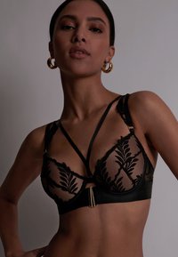 Soutien-gorge en dentelle noire avec tissu transparent, présentant des motifs de feuilles, des bretelles réglables et un accent en métal doré au centre devant.