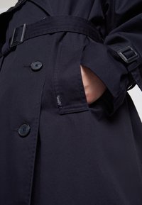 Trench coat blu navy realizzato in tessuto resistente, con grandi bottoni neri, una cintura e tasche laterali. Mostra i dettagli delle tasche e delle cuciture.