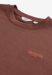 Ruskea huppari, jossa on pyöreä pääntie, rento malli ja oranssilla brodeerattu "AMSTERDAM DAMSTRAAT" -logo. Pehmeä, tekstuuri-kangas.