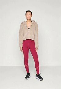 Fleece zip-up hoodie i beige, parret med burgunder leggins med mesh-paneler, og sorte sportssko med hvide detaljer.