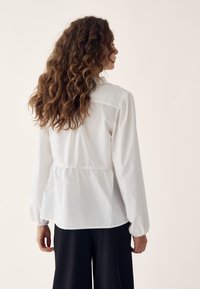 Woman with long curly hair wearing a white long-sleeve blouse and black wide-leg pants, shown from the back.