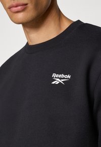 Czarna bluza z miękkiego materiału z prążkowanym kołnierzem, z białym logo Reebok na piersi. Zbliżenie podkreślające teksturę.