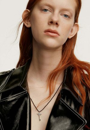 Chaqueta de piel sintética negra con costuras blancas, superpuesta con un collar de doble cadena que presenta un colgante de llave de plata. Se pueden ver cabellos rojos.