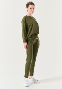 Olijfgroene loungewear set bestaande uit een losse top met lange mouwen en joggers met een trekkoord. Gecombineerd met witte sneakers. Soepele textuur.