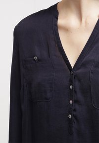 Marinblus i lättviktsmaterial med V-ringning, knappstängning och två bröstfickor med knappar. Slät yta.