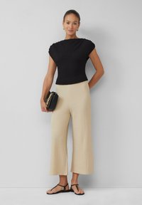 Zwart textuur top met kapmouwen, gecombineerd met beige wijde broek met een subtiel patroon. Model houdt een zwarte handtas vast en draagt sandalen.