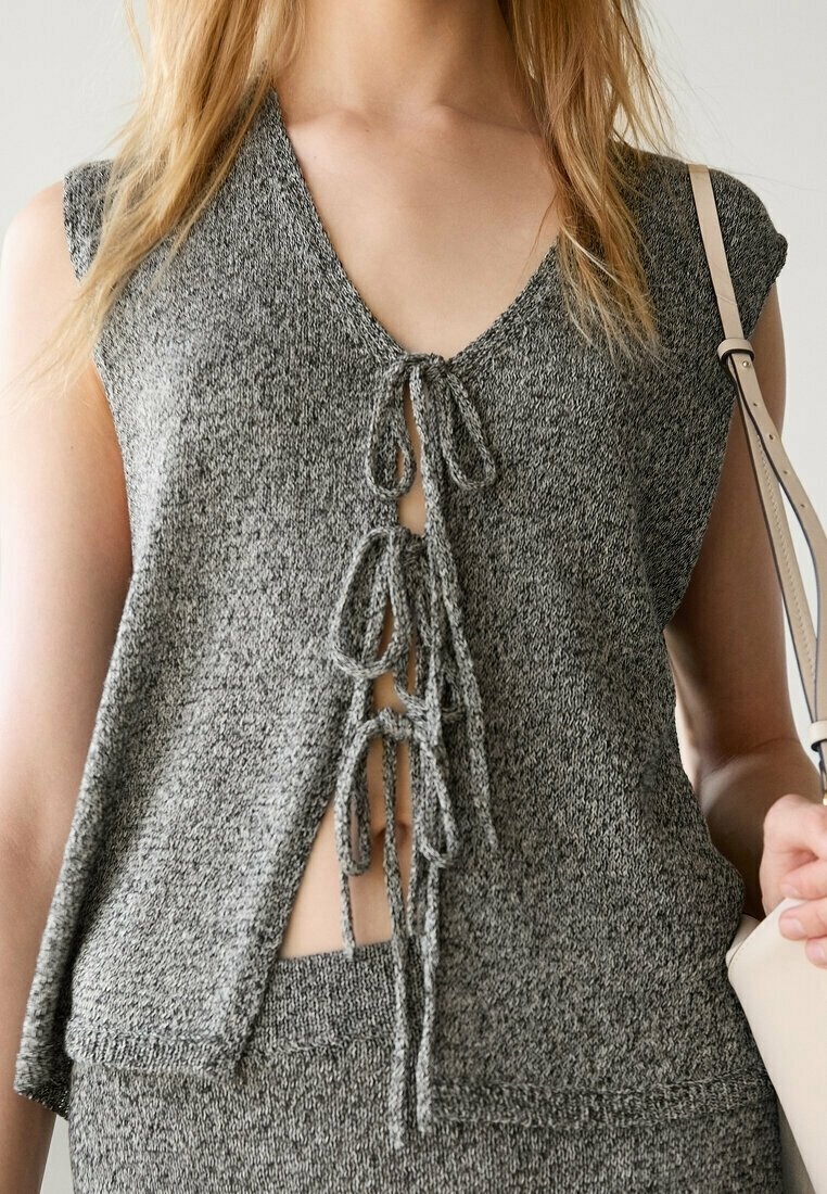 Femme portant un débardeur gris en maille avec trois nœuds noués sur le devant, assorti à un pantalon en maille assorti et tenant un sac beige.