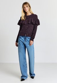 Dunkellila, strukturierter Cardigan mit Rüschensäumen und Knopfleiste, kombiniert mit hellblauen, weit geschnittenen Jeans und schwarzen, spitzen Schuhen.