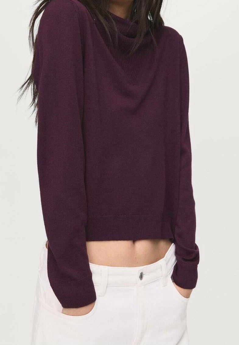 Femme portant un pull court à manches longues violet foncé et un pantalon blanc, les mains dans les poches, debout devant un fond uni.