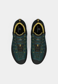 Športové topánky v tmavozelenej semišovej koži s textúrovanou čiernou gumovou podrážkou. Obsahujú žlté akcenty, hnedé šnúrky a sieťovanú jazyk pre priedušnosť.