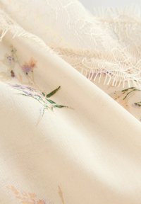 Tissu crème avec de délicats motifs floraux dans des couleurs pastel, agrémenté d'une bordure en dentelle ajoutant texture et détails sur les bords.