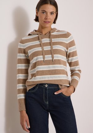 Femme portant un pull à capuche rayé beige, blanc et gris avec des cordons et un jean bleu foncé, une main dans la poche.