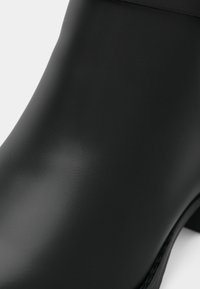 Tuvumā redzam melnu, gludu ādas potīšu zābaku ar zemu papēdi uz baltā fona.