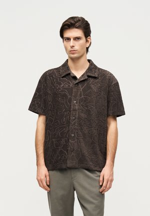 Sötét hajú fiatal férfi rövid ujjú, gombos, texturált barna inget visel absztrakt mintákkal és világosszürke nadrágot.