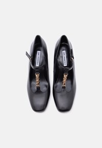 Γυναικεία παπούτσια από μαύρο δέρμα με T-λαιμόκοψη και χρυσά γράμματα "KARL" ως γούρια, τετράγωνη μύτη και κούμπωμα με αγκράφα, απεικονιζόμενα από πάνω σε λευκό φόντο.