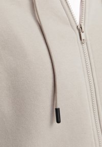 Hellbeige Zip-Up Hoodie mit weicher Textur, ausgestattet mit einer kontrastierenden schwarzen Spitze am Kordelzug und standardmäßigem Frontreißverschluss.