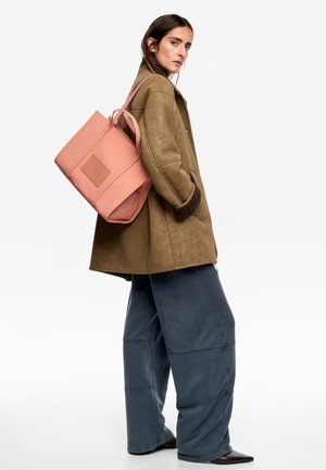 Abrigo marrón de ante sintético, pantalones azules holgados y un bolso tote rosa. El bolso tote tiene forma rectangular, asas superiores y un parche con logo.